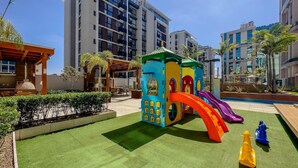 Zona de juegos infantiles con un tobogán, rodeada de follaje y edificios modernos.