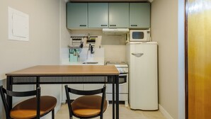 A compact kitchen with teal cabinets, a white refrigerator, and a microwave. There is a wooden table with two chairs. - Apto 1Q 350m do PQ. Bosque do Sudoeste - SVS0219 (Sudoeste e Octogonal)