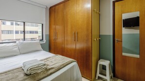 A bedroom with a bed, a wardrobe, a small stool, and a TV mounted on the wall. - Apto 1Q 350m do PQ. Bosque do Sudoeste - SVS0219 (Sudoeste e Octogonal)