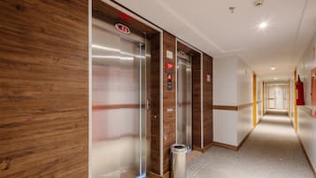 Pasillo de hotel con dos ascensores, paneles de madera en las paredes, un extintor y un cubo de basura.