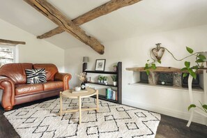 A cozy living room with a brown leather sofa, a small wooden side table, a white patterned rug, a bookshelf, and a wooden shelf with plants. - Jackson s Barn in Water Yeat (Ulverston)