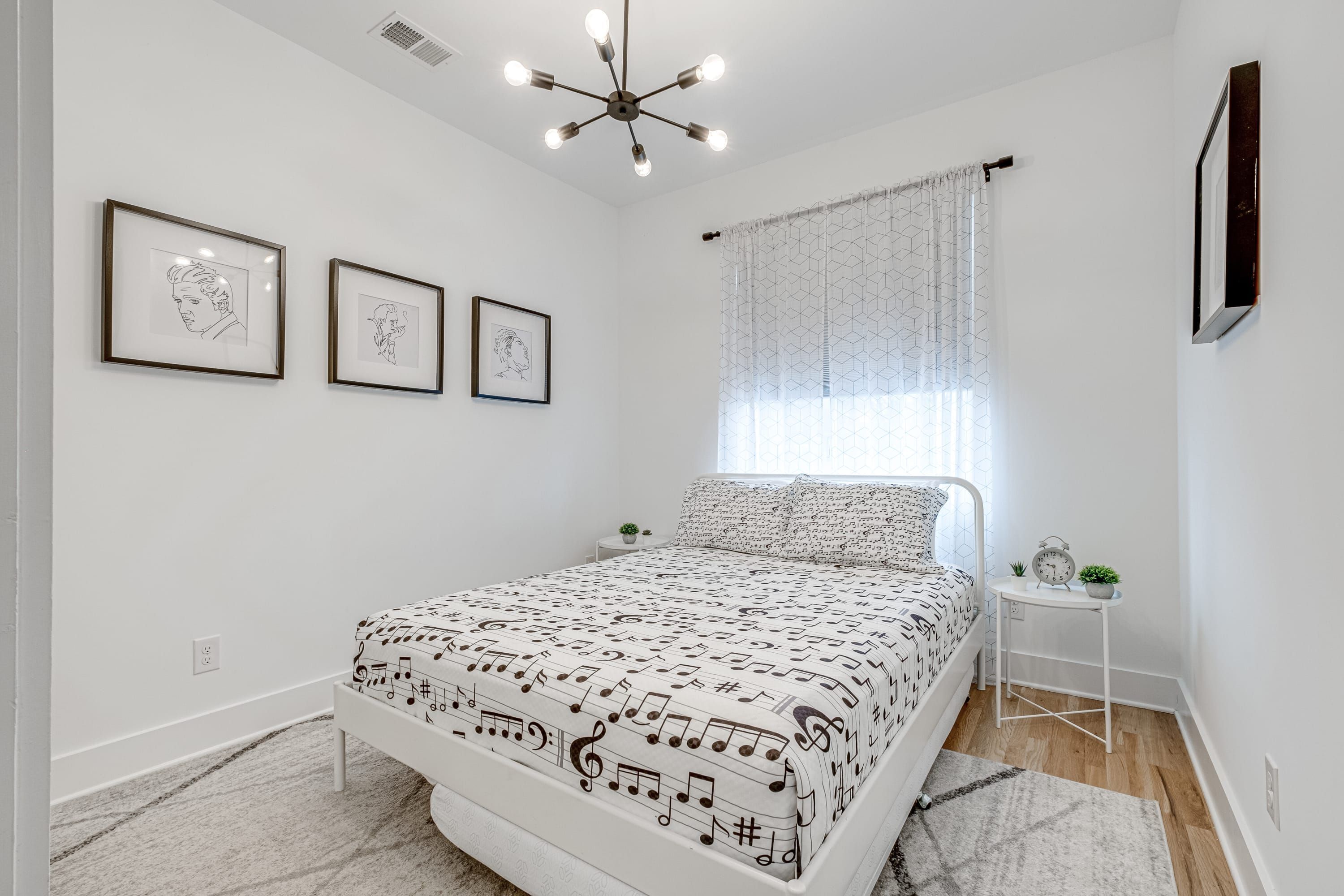 A bedroom with a bed, framed artwork on the wall, a small bedside table with a plant, and a window with a curtain.