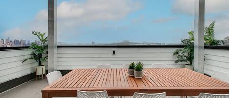 Ein Holztisch mit weißen Stühlen auf einer Dachterrasse, Blick auf eine Stadtlandschaft.