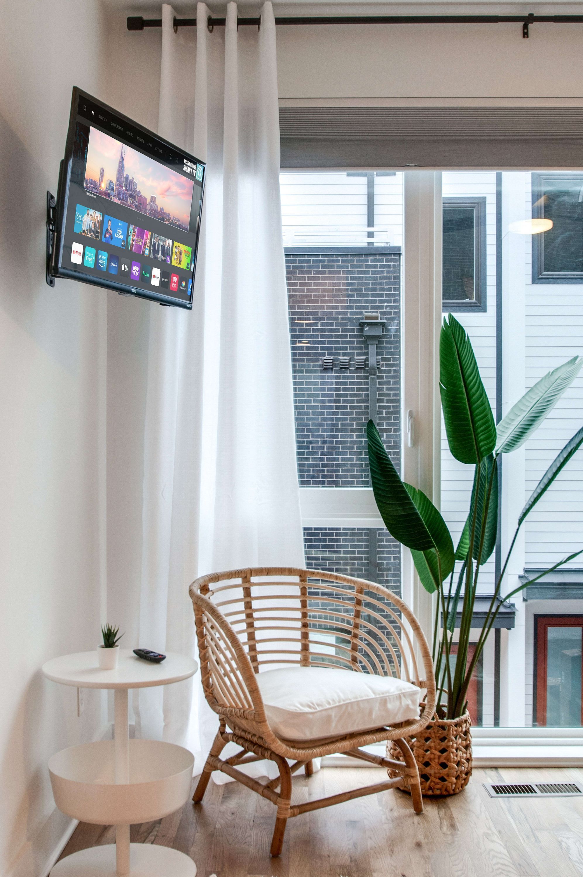 Een kamer met een groot raam, een rieten stoel, een klein tafeltje en een tv met een stadsbeeld erop.