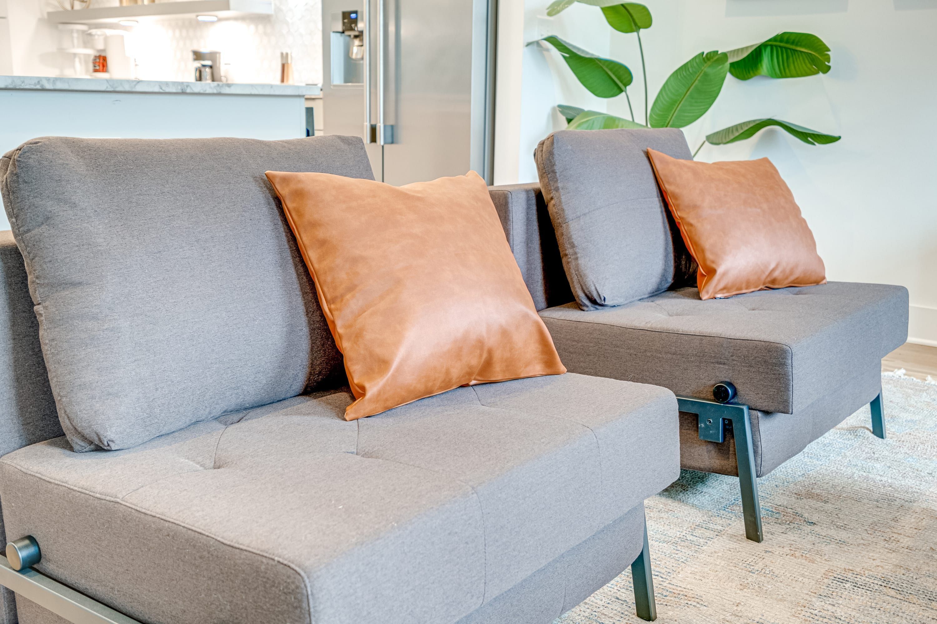 Een grijze bank met twee bruine kussens en een klein lade, geplaatst in een moderne woonkamer met een grote plant en een keuken op de achtergrond.