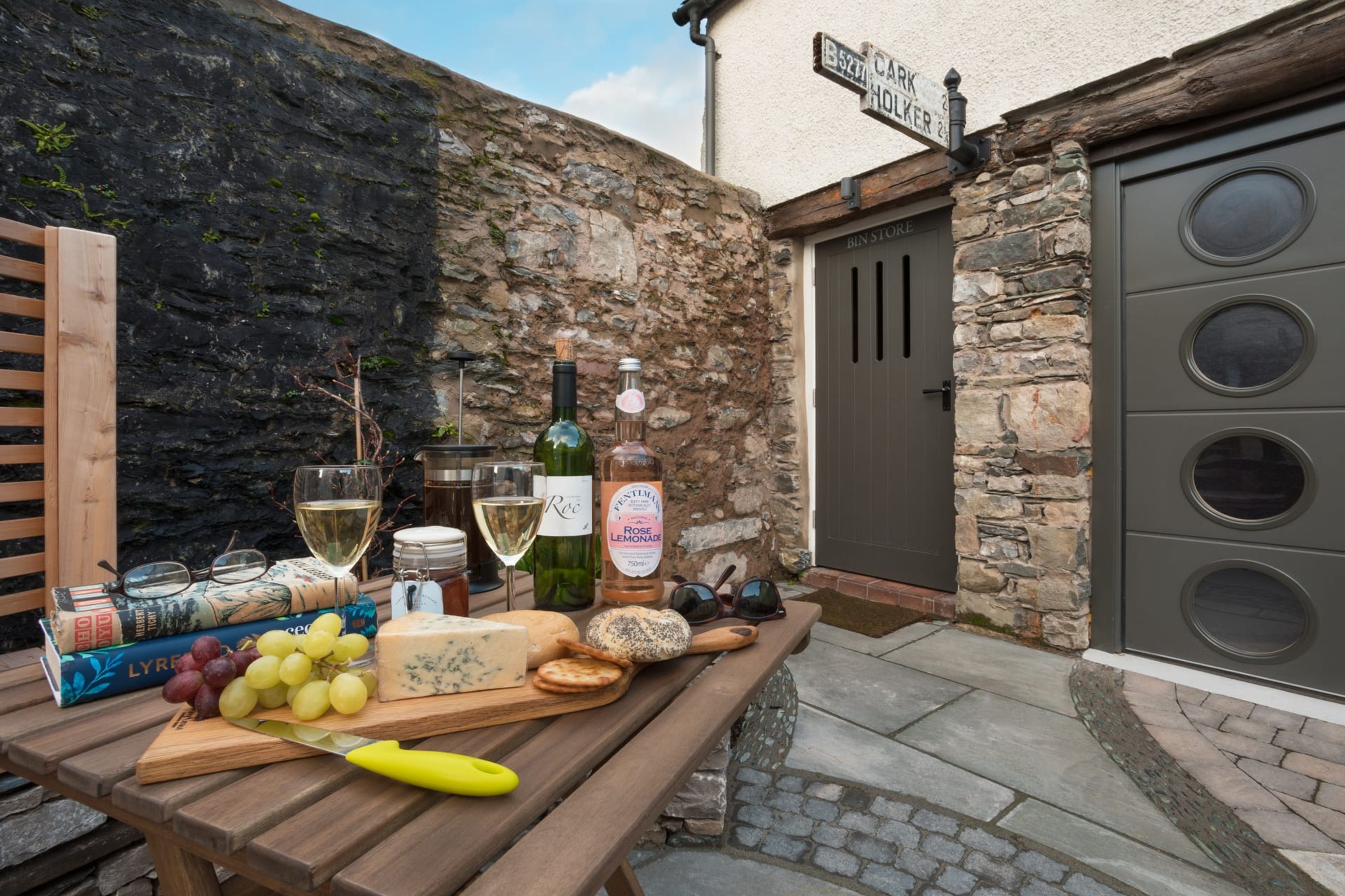 A wooden table with a cheese board, wine glasses, and books in an outdoor setting with stone walls and a garage door.