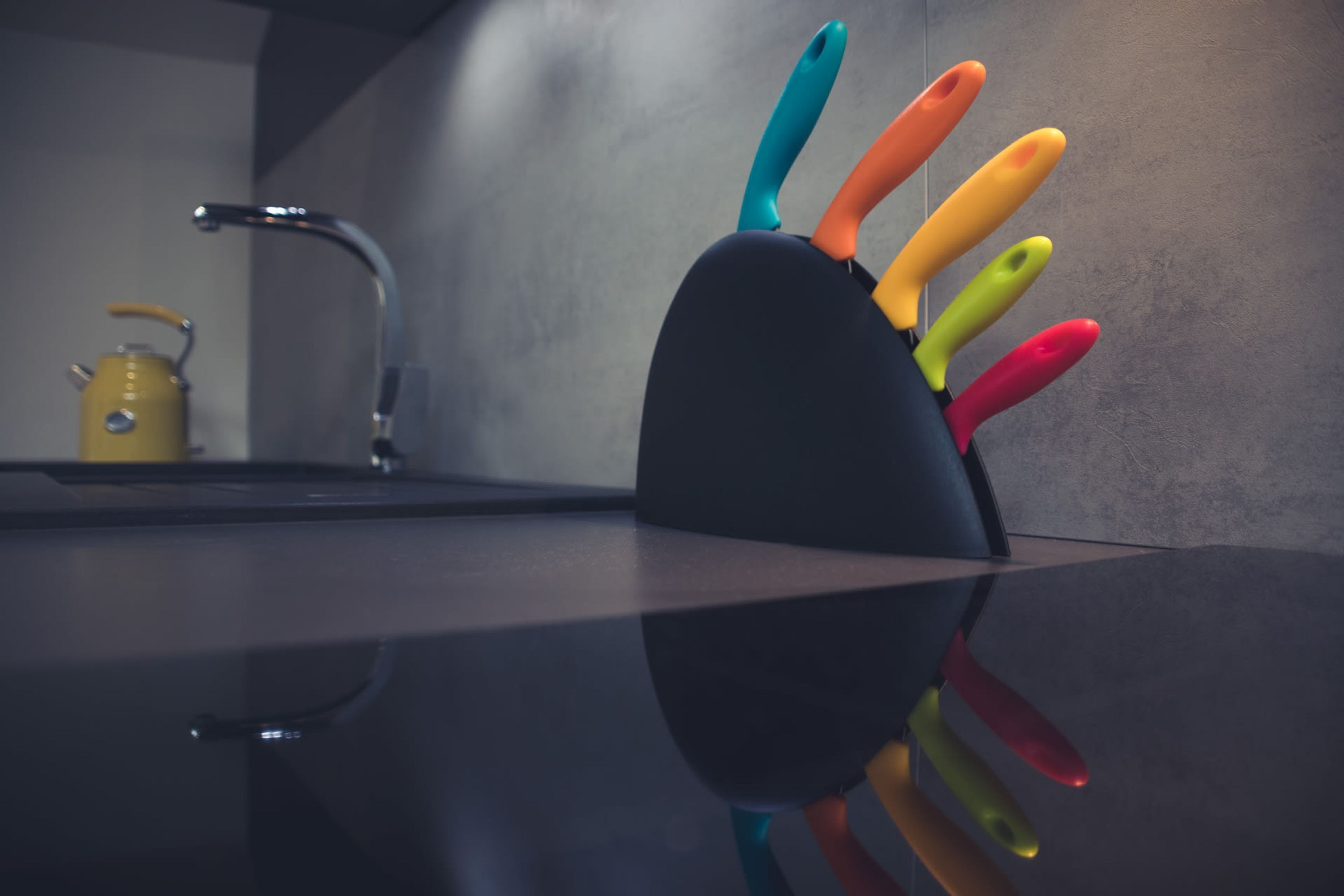 A kitchen counter with a knife block holding colorful knives and a yellow kettle in the background.