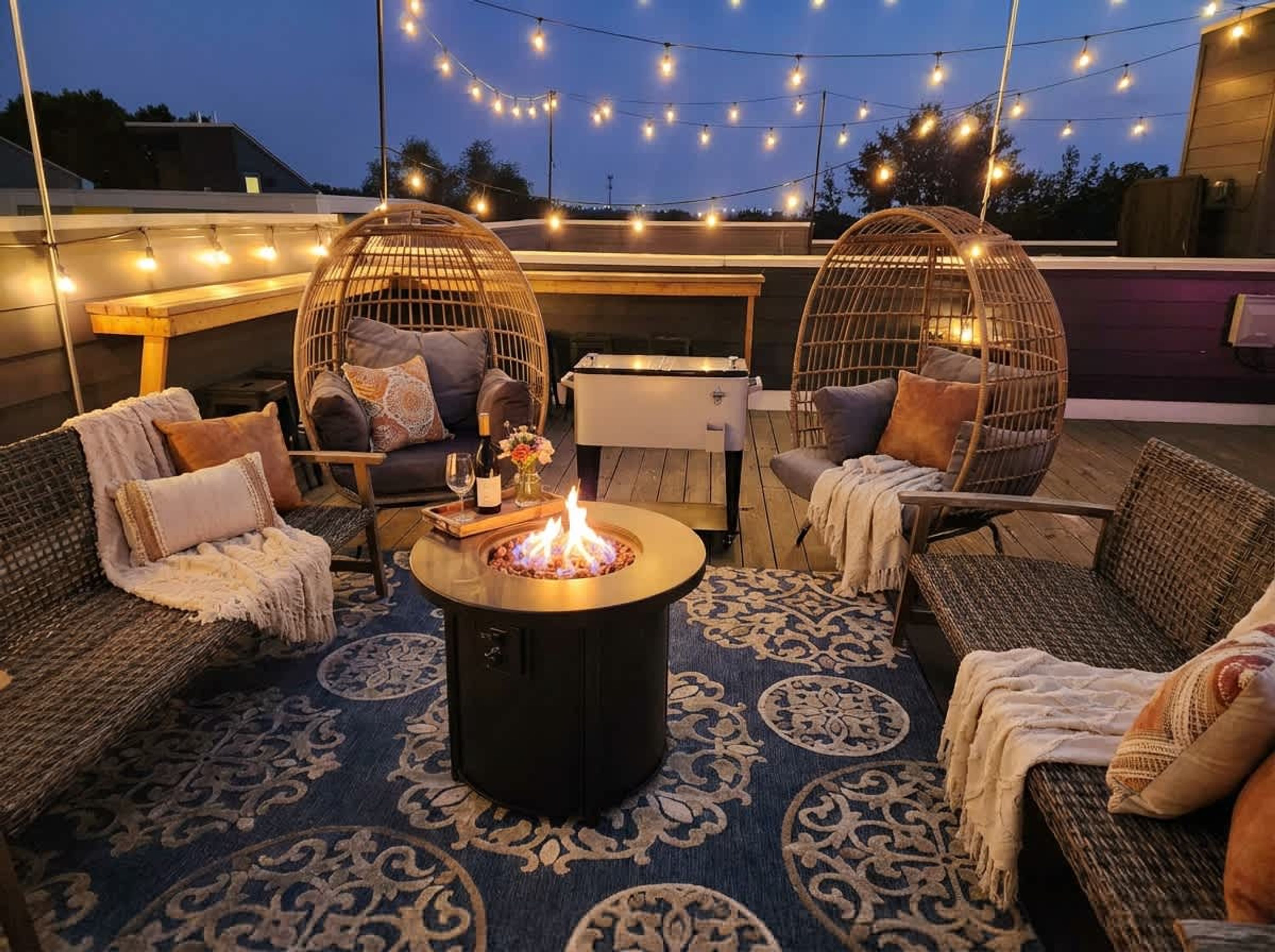 An outdoor seating area with wicker furniture, a central fire pit, and string lights.