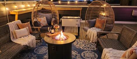 An outdoor seating area with wicker furniture, a central fire pit, and string lights.
