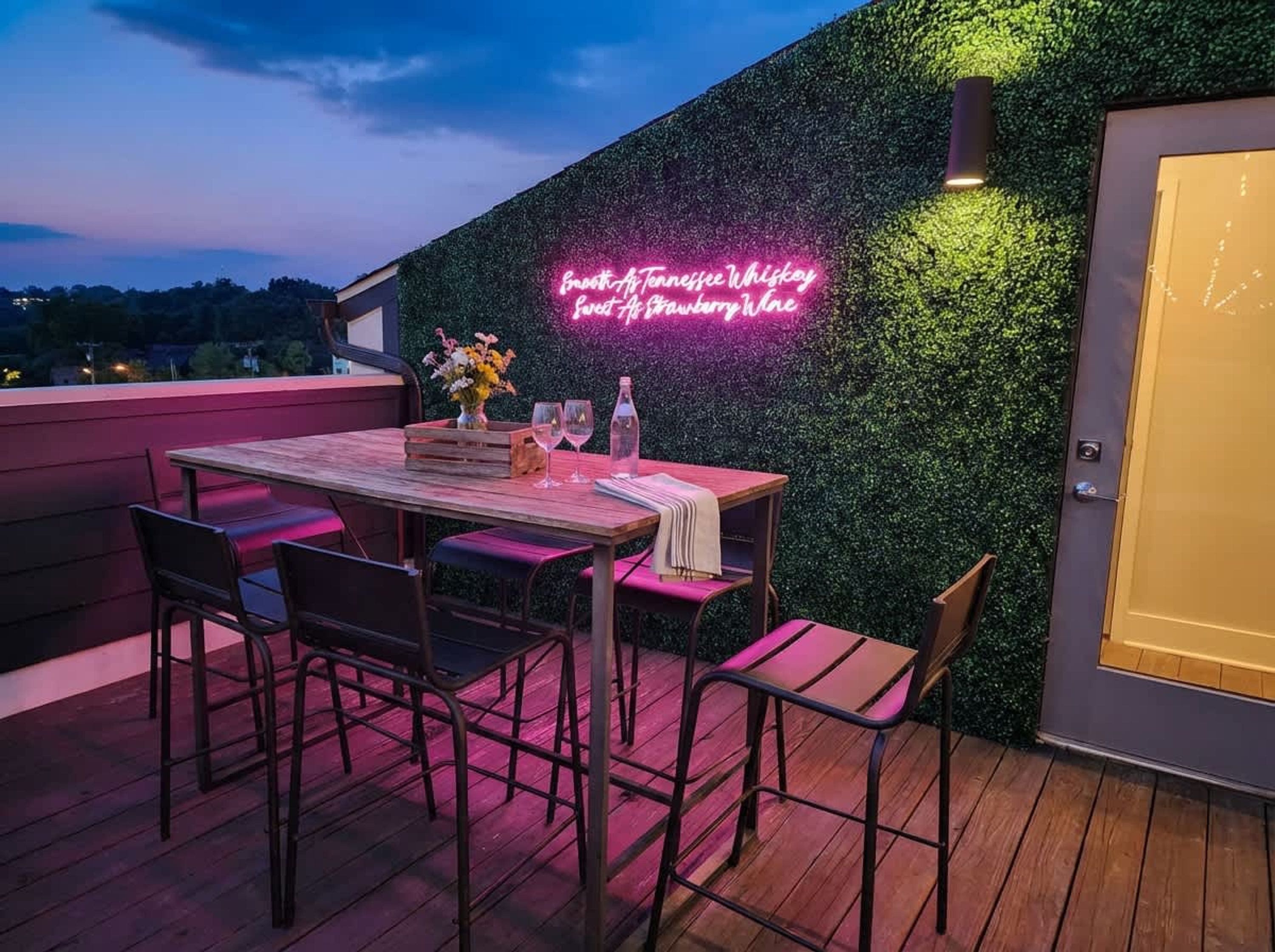 A rooftop patio with a neon sign, wooden table, chairs, and a decorative vase.