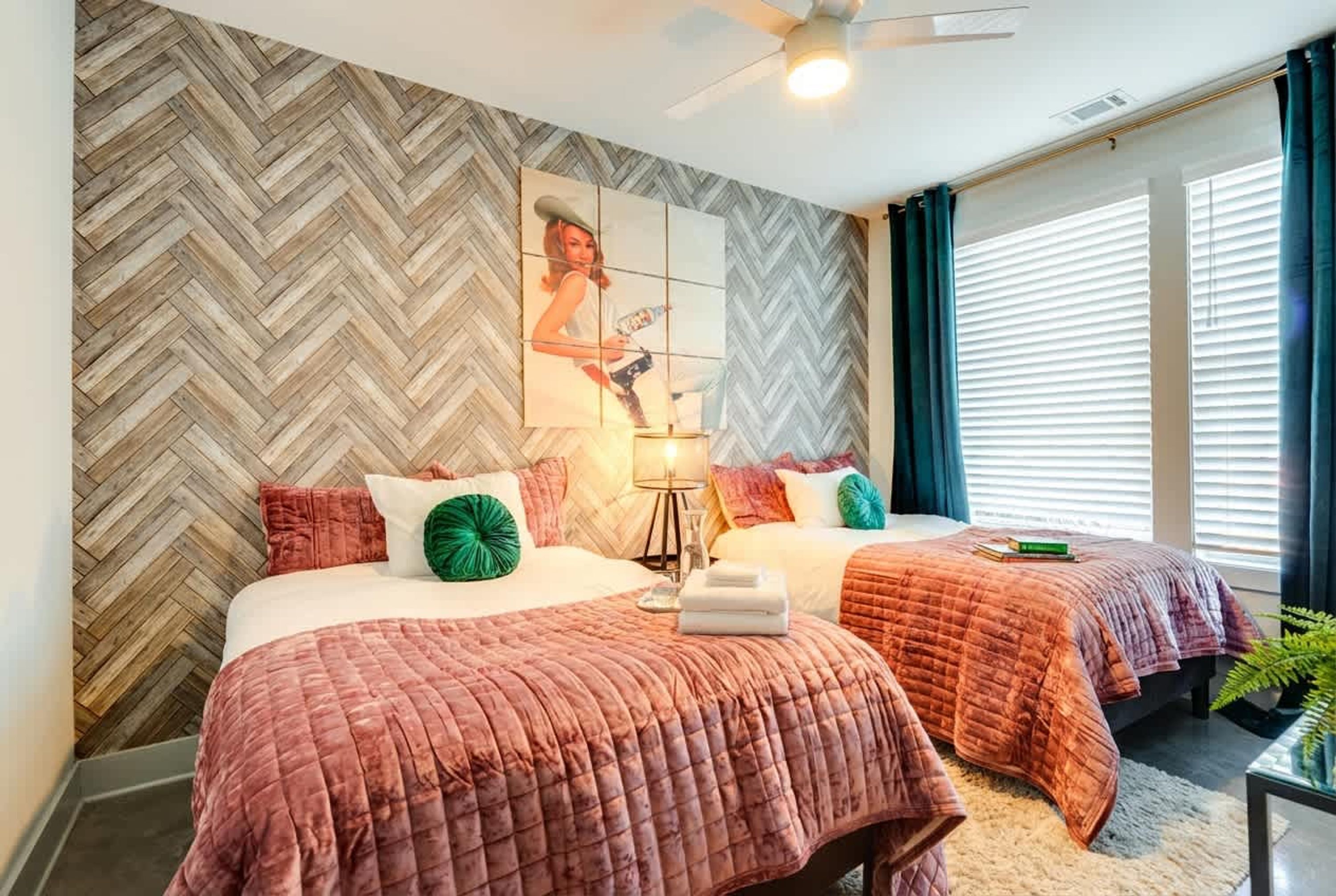 A hotel room with two beds, a herringbone-patterned wall, a ceiling fan, and a window with blinds.