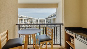 A balcony with a round table and two chairs, a kitchen area with a black countertop, and a view of buildings in the distance. - Condomínio Bem localizado com Piscina TCR (Taguatinga)
