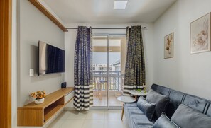 Ein Wohnzimmer mit einer Couch, einem Fernseher, einer Holzkonsole und einem Balkon mit Vorhängen.