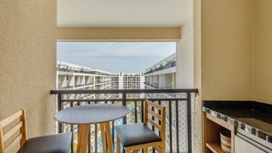 A balcony with a round table and two chairs, a kitchen area with a black countertop, and a view of buildings in the distance. - Condomínio Bem localizado com Piscina TCR (Taguatinga)