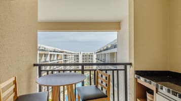 Uma varanda com uma mesa redonda e duas cadeiras, uma área de cozinha com bancada preta e vista para edifícios ao longe.
