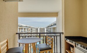 Un balcón con una mesa redonda y dos sillas, una zona de cocina con encimera negra, y vistas a edificios distantes.