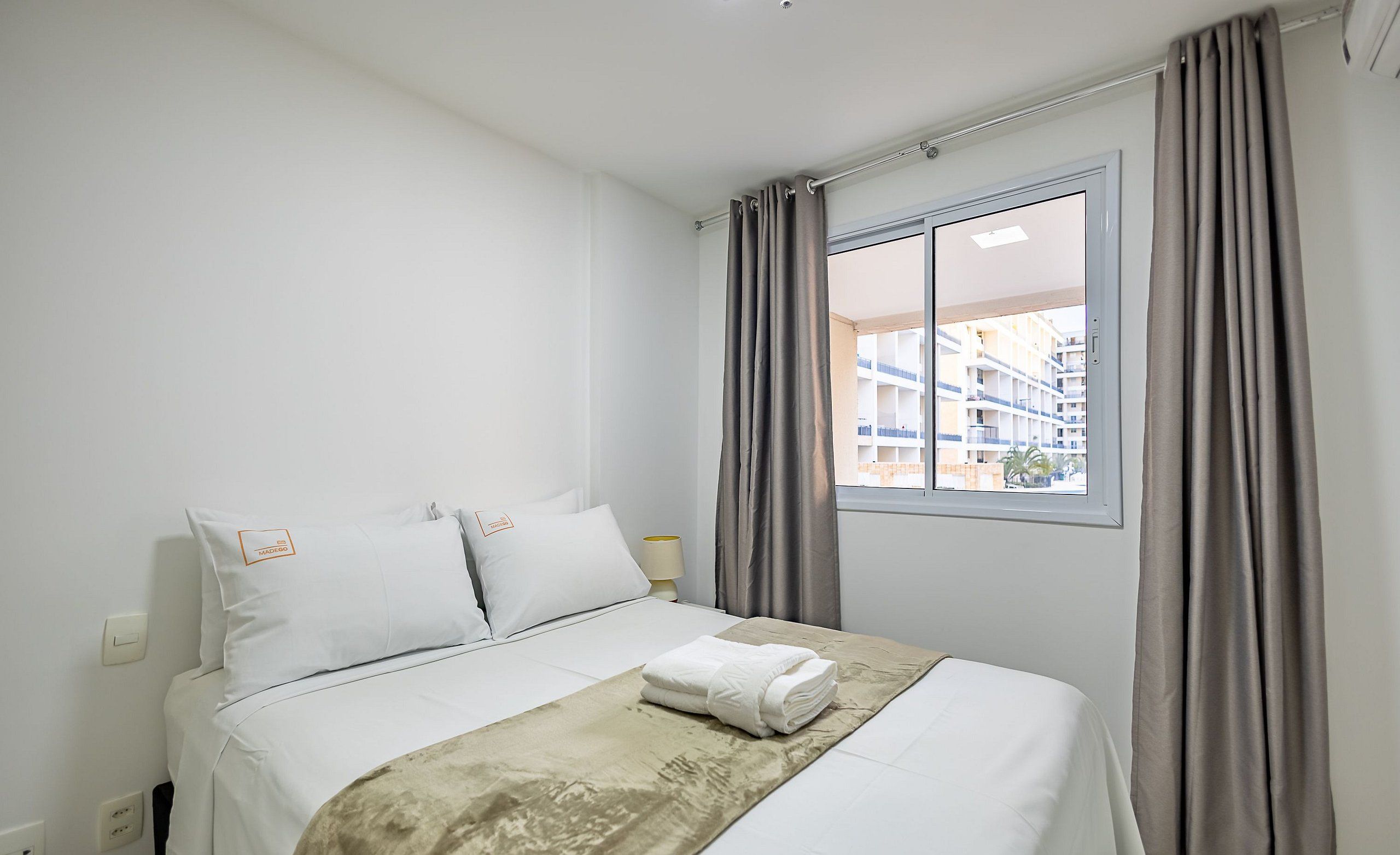 Una cama bien hecha con edredón blanco y manta color beige, una mesita de noche con lámpara, y una ventana con cortinas.