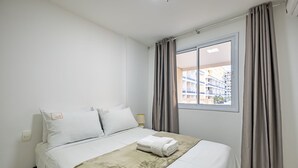 A neatly made bed with a white comforter and a beige blanket, a nightstand with a lamp, and a window with curtains. - Condomínio Bem localizado com Piscina TCR (Taguatinga)