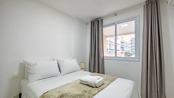 Una cama bien hecha con edredón blanco y manta color beige, una mesita de noche con lámpara, y una ventana con cortinas.