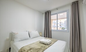 Una cama bien hecha con edredón blanco y manta color beige, una mesita de noche con lámpara, y una ventana con cortinas.