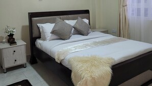 A neatly made bed with a fluffy white throw, a bedside table with a vase of flowers, and a dark wooden headboard. - Kingdom Corner Suites (Nairobi)