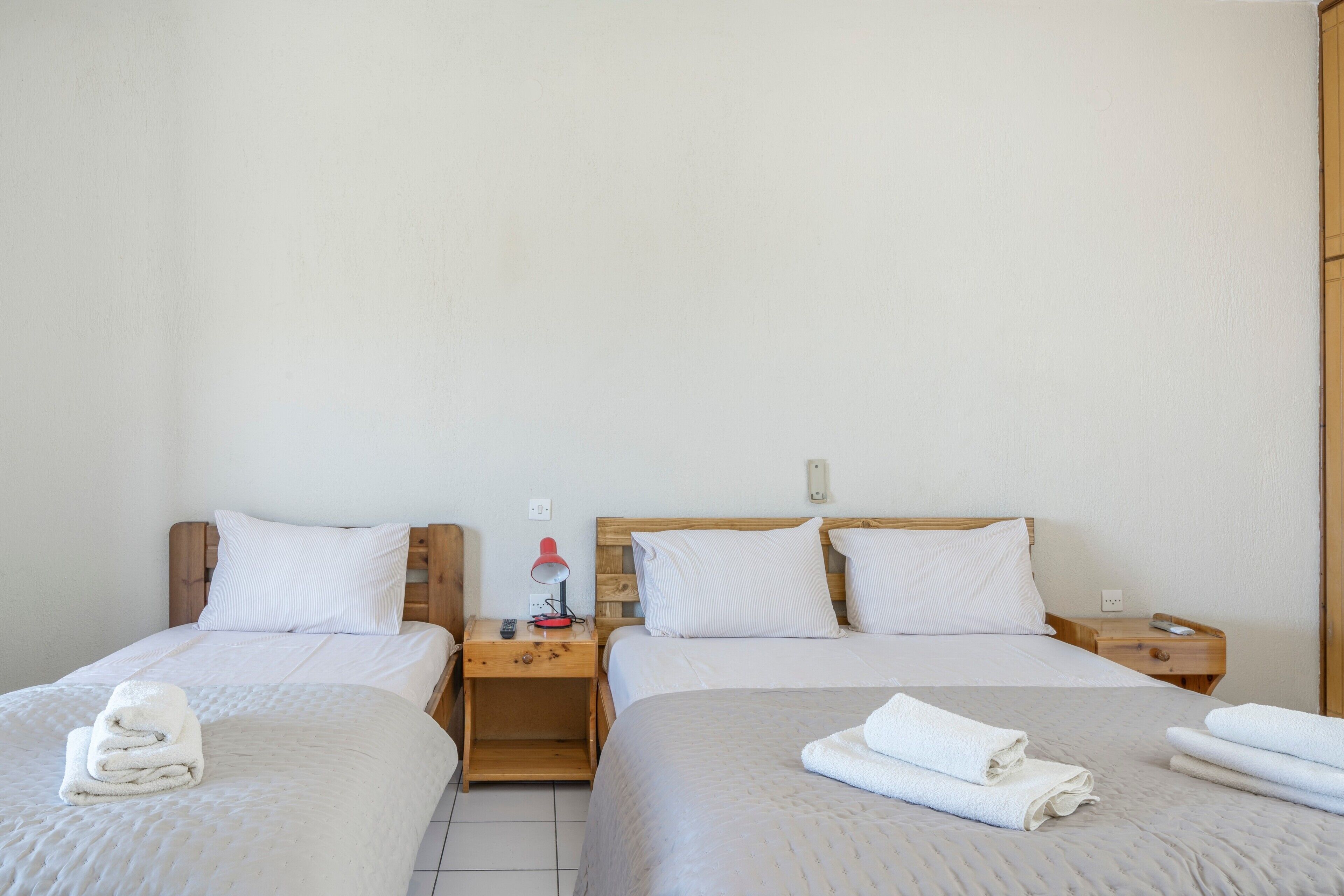 A hotel room with two beds, wooden headboards, white bedding, and a nightstand with a lamp and remote control.