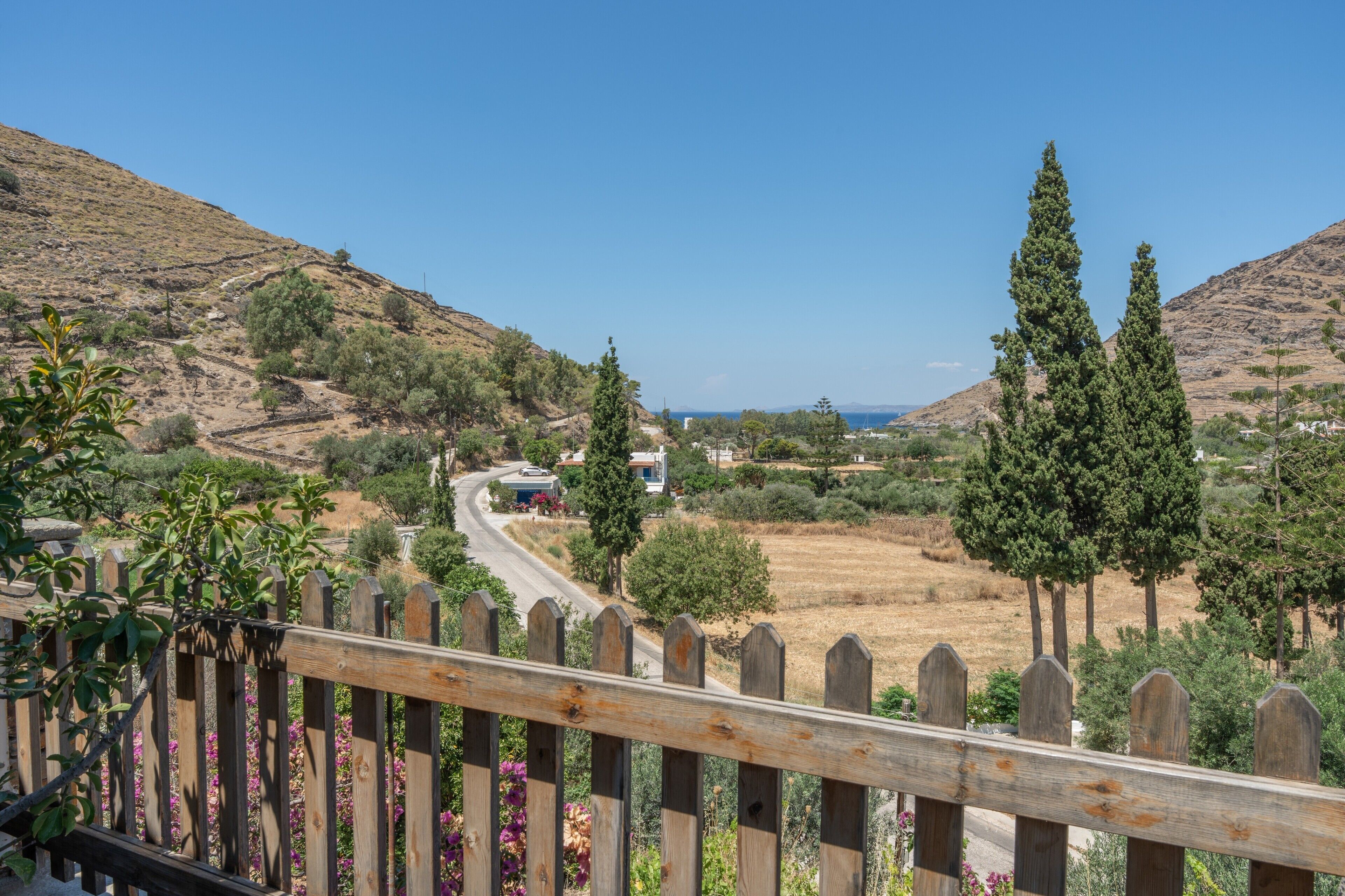 A rural landscape with a winding road, a wooden fence, and a view of a valley with trees and buildings.