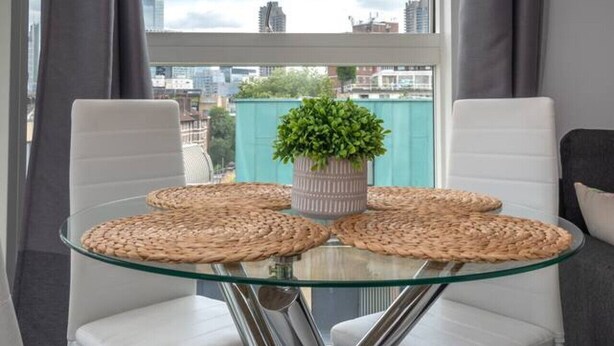 A modern dining area with a glass table surrounded by white chairs, a potted plant, and woven placemats. - Clerkenwell Flat Near Old Street Pet Friendly (London)