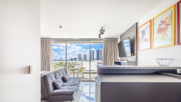 A modern living room with a grey sofa, a flat-screen TV mounted on the wall, and a city view through large windows. - Complete Stay and Pool in Aguas Claras - DPA (Águas Claras)