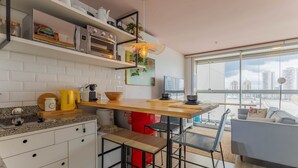 A modern kitchen with white cabinets, a wooden countertop, and a red refrigerator. There is a dining area with a table and chairs, and a large window offering a city view. - Complete Stay and Pool in Aguas Claras - DPA (Águas Claras)