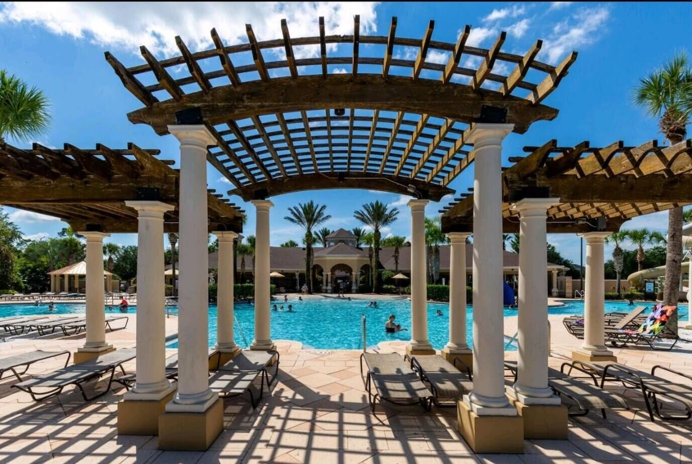 A pool area with a pergola structure, lounge chairs, and palm trees in the background.