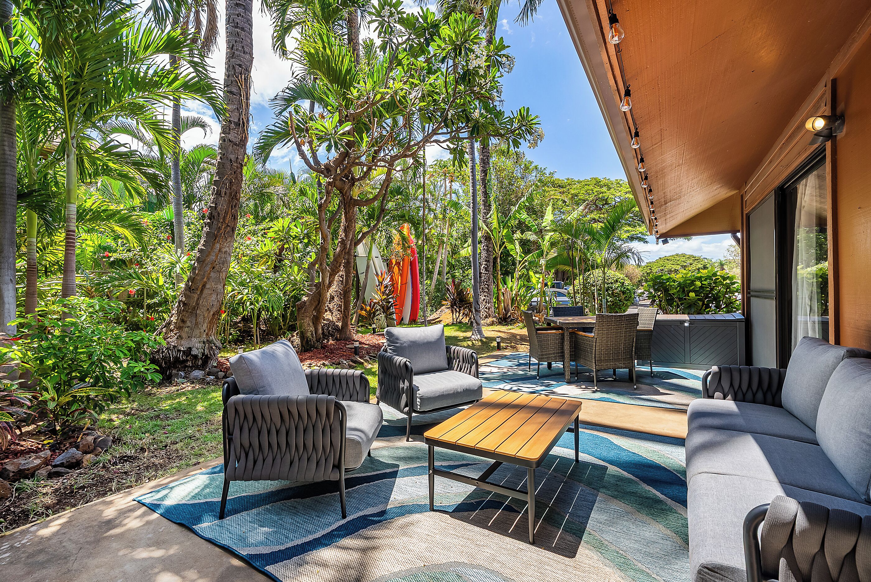 An outdoor patio with a sofa, chairs, and a table, surrounded by lush greenery and palm trees.