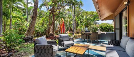An outdoor patio with a sofa, chairs, and a table, surrounded by lush greenery and palm trees.