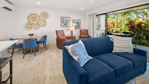 A modern living room with a blue sofa, two armchairs, a dining table with chairs, a ceiling fan, and a view of a garden through a glass door.