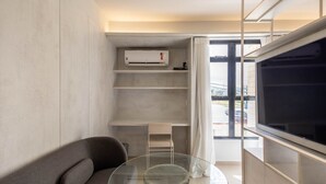 A modern living room with a grey sofa, a glass coffee table, a mounted air conditioner, and a large window with a view of the outside. - Studio Moderno prox. do Shoppping Iguatemi SHL0105 (Lago Norte)