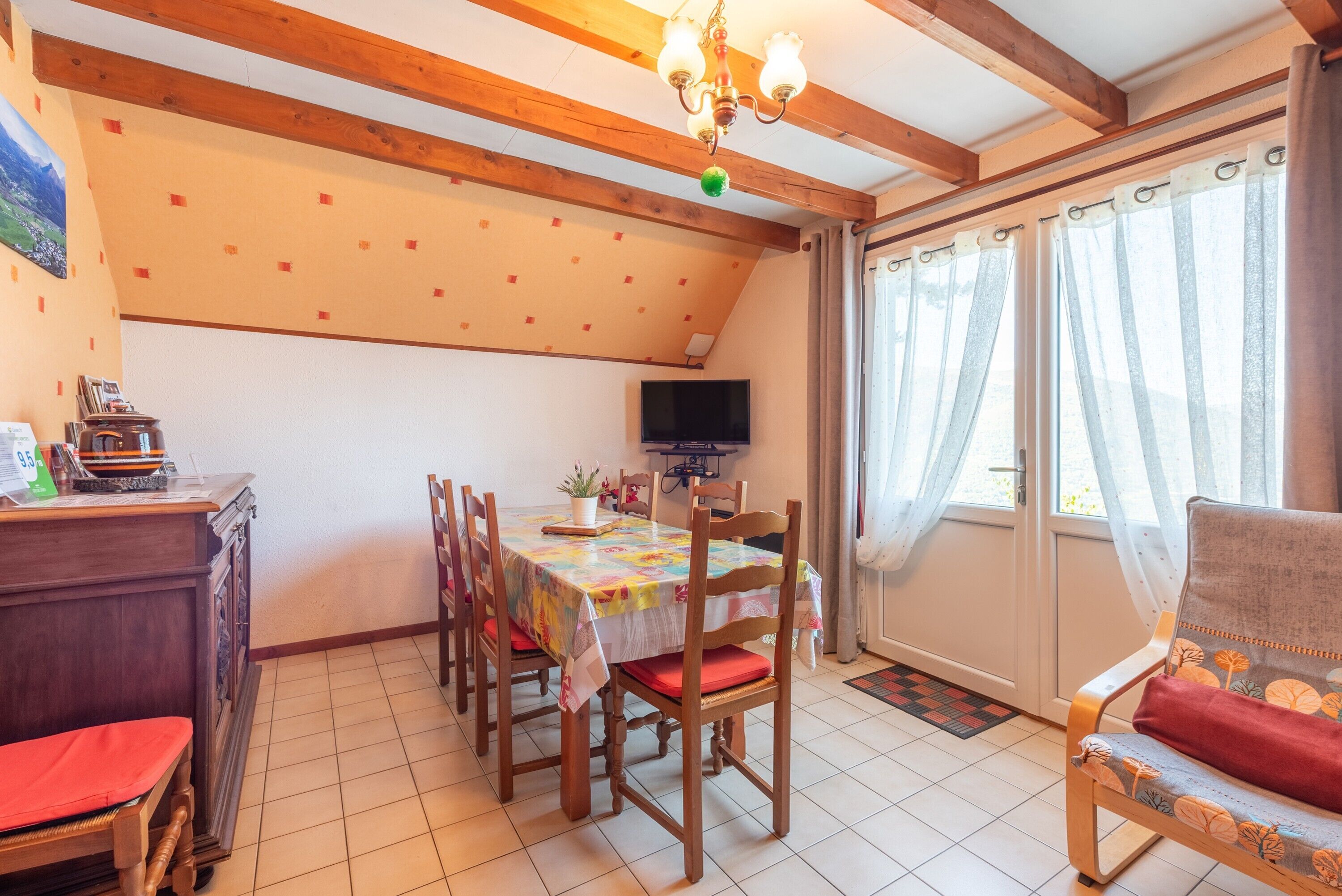 Un comedor con mesa, sillas, televisor y ventana con cortinas.