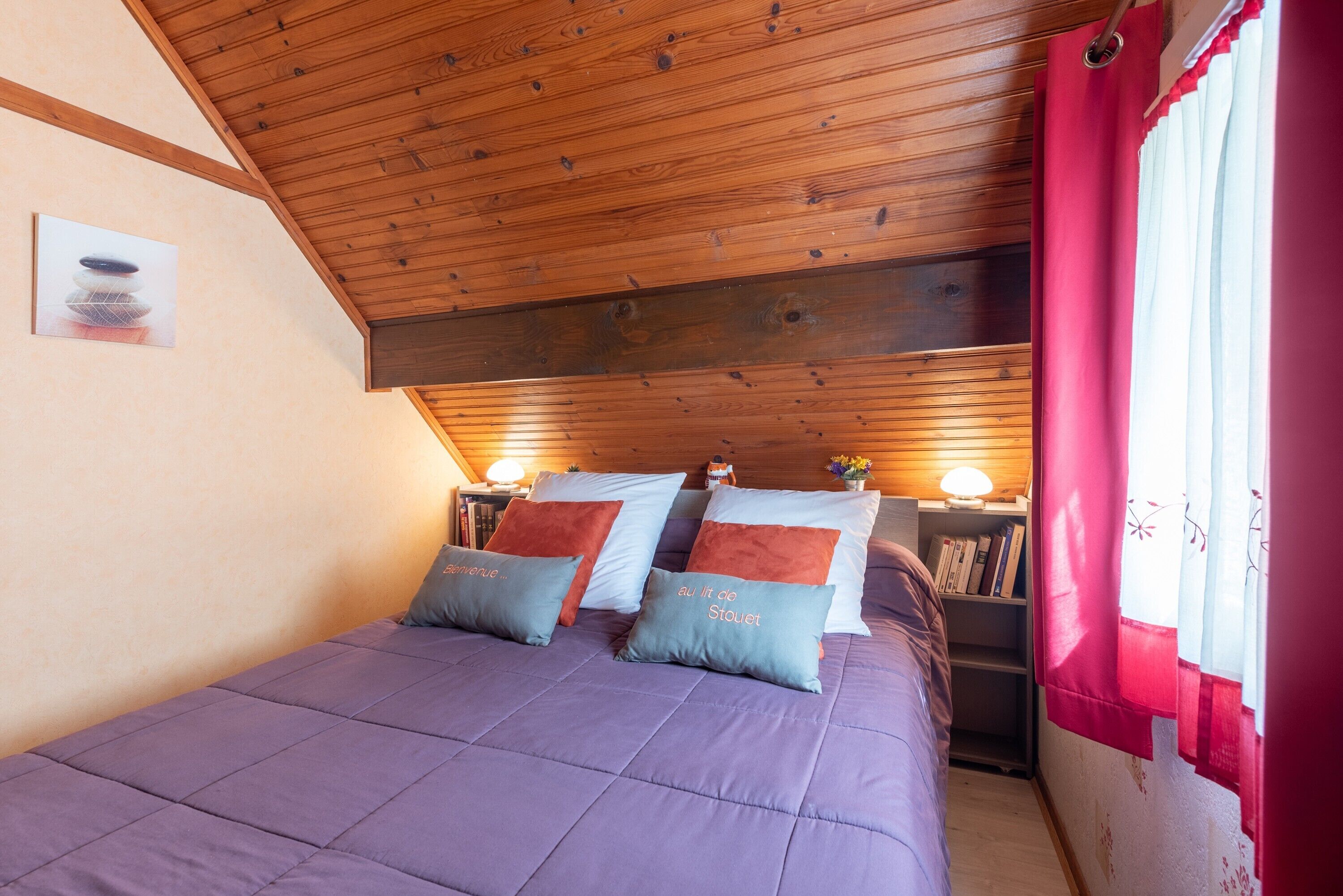 A bedroom with a wooden ceiling, a bed with purple bedding, and a nightstand with a lamp.