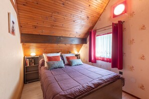 A bedroom with a wooden ceiling, a bed with a purple bedspread, two bedside tables with lamps, and a window with red curtains.