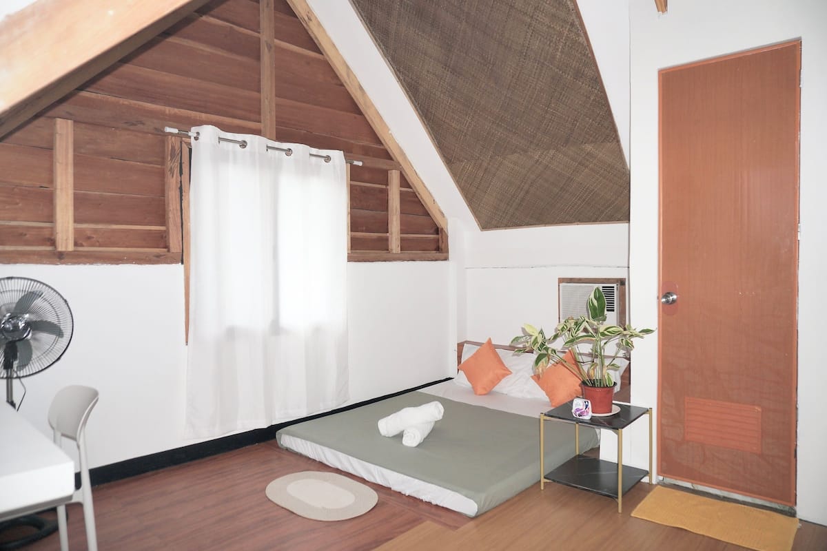 Een kleine, goed verlichte kamer met een enkel bed, een bureau, een stoel, een ventilator en een deur.