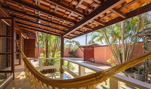 A hammock on a balcony with a view of a garden and a building with a tiled roof. - Charming House w/ BBQ in Vila Planalto Act029 (Brasília)