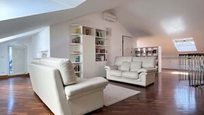 A spacious living room with a high ceiling, a white sofa, a white armchair, and a built-in bookshelf.
