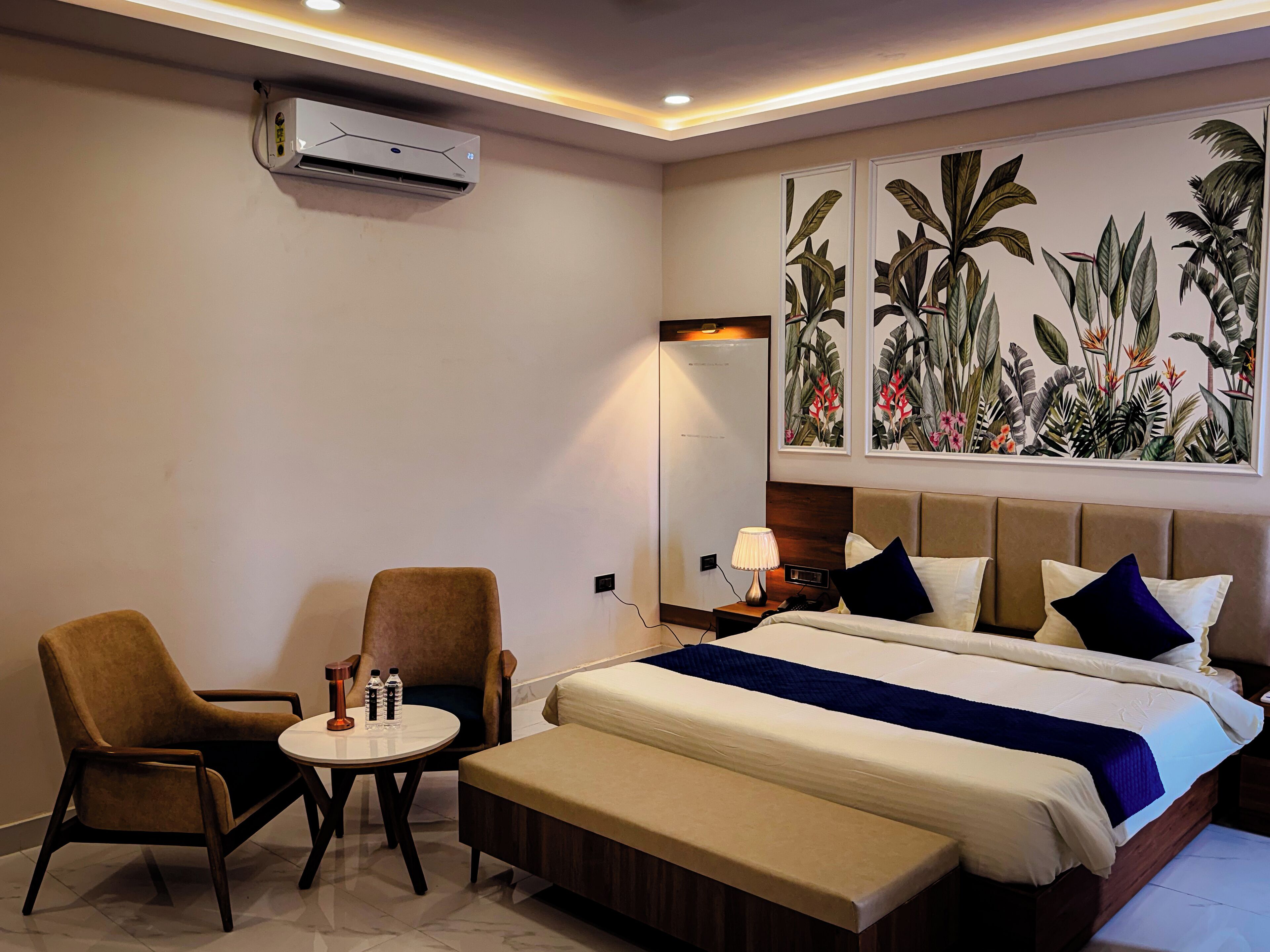 A hotel room with a bed, a bench, two chairs, a small table, an air conditioning unit, and a wall-mounted mirror.