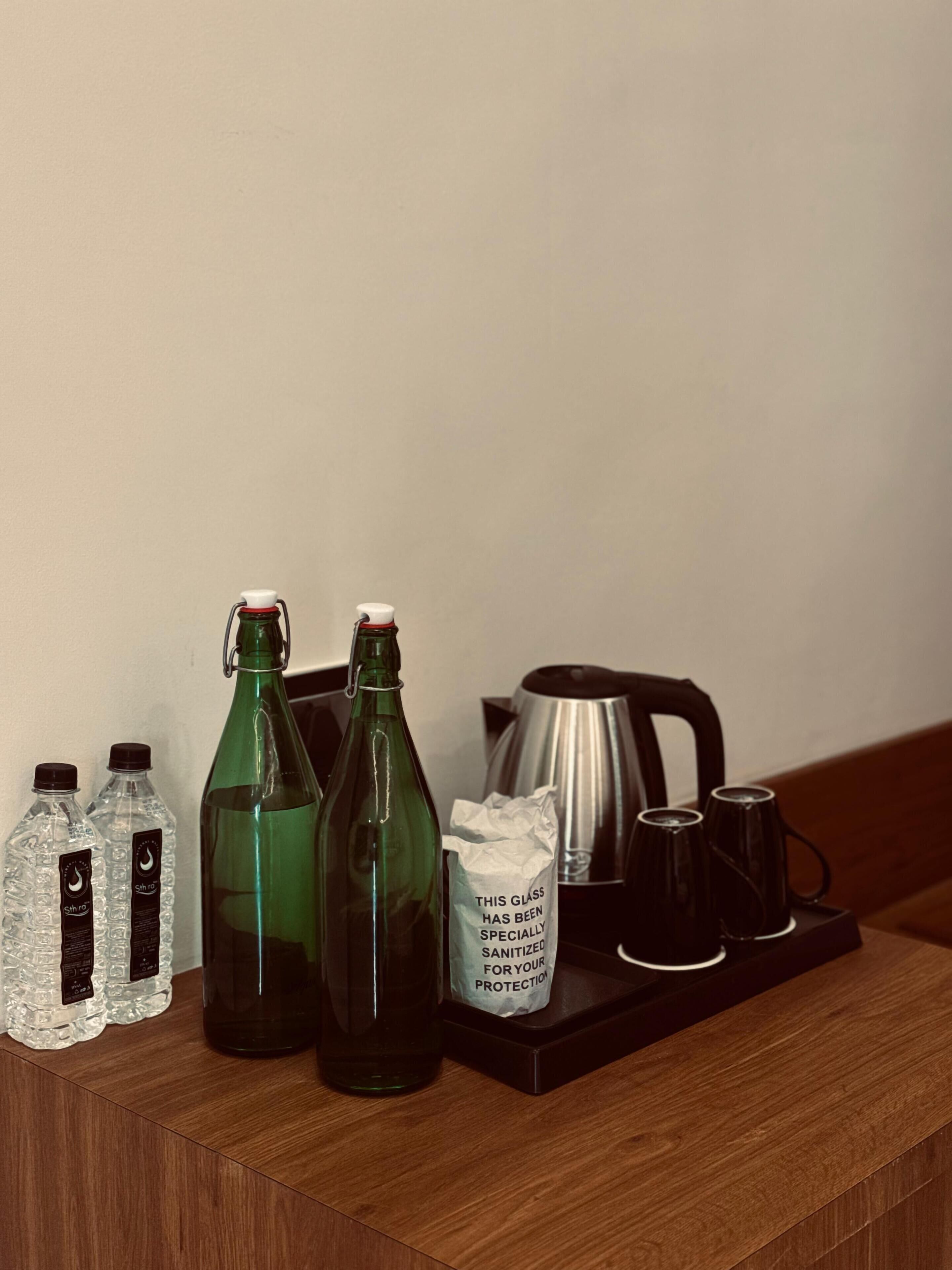 A wooden table with a glass of water, two bottles of water, a green bottle, a stainless steel coffee pot, and a tray with a coffee maker and cups.