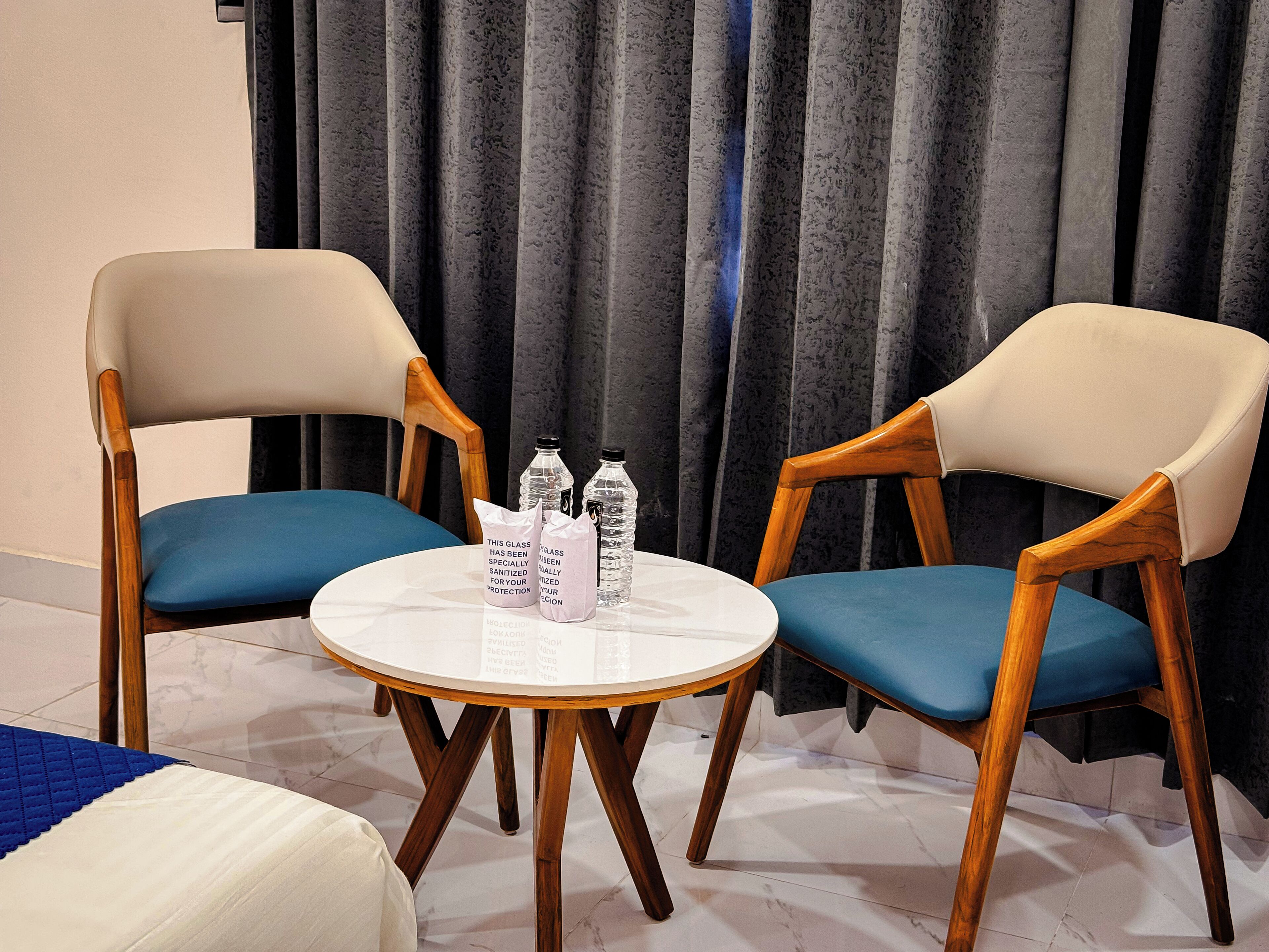 Ein Zimmer mit zwei Holzsesseln mit blauen Kissen, einem kleinen runden Tisch mit weißer Tischplatte und zwei Wasserflaschen.