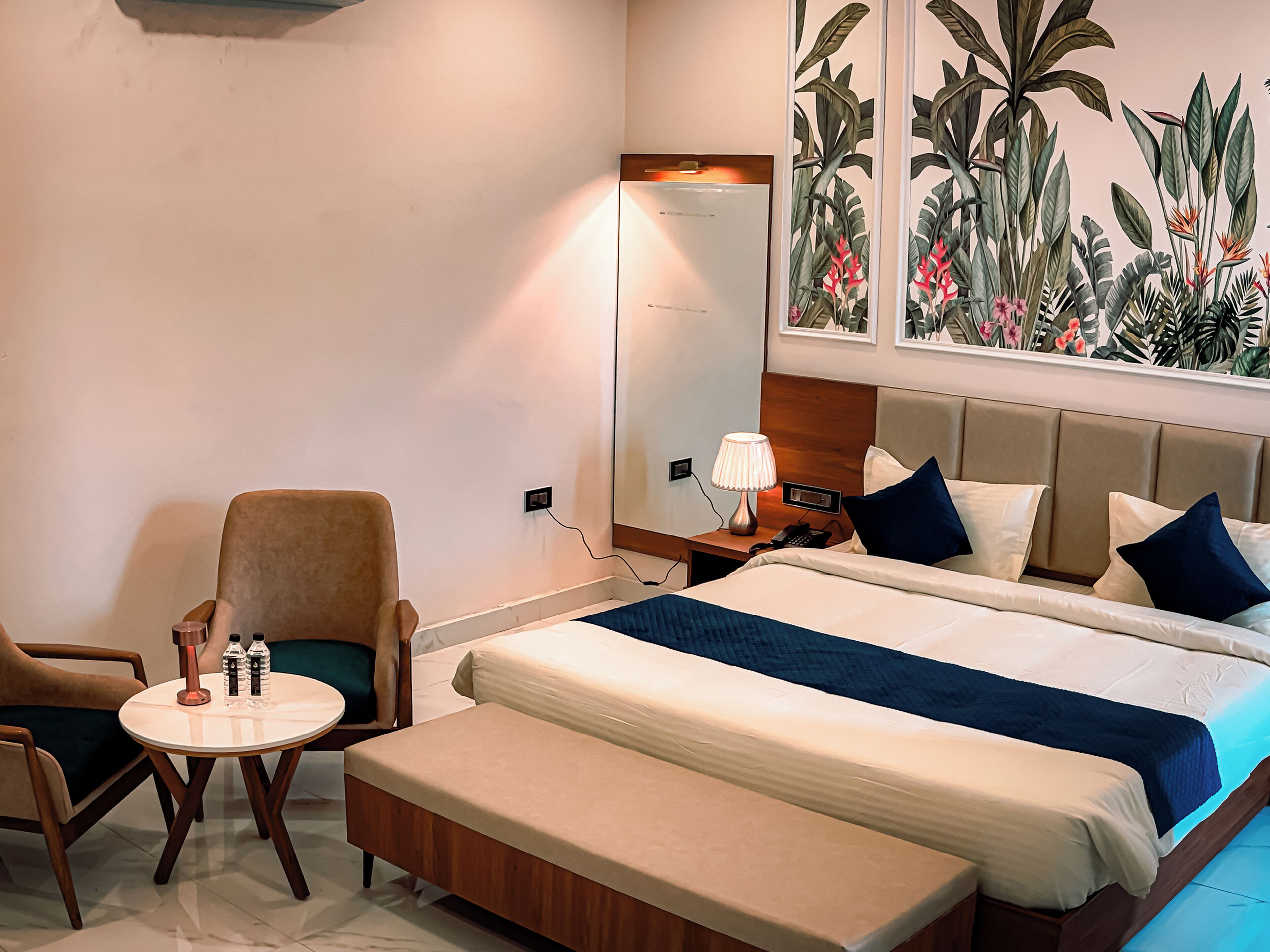 Hotelkamer met een bed, een stoel, een tafeltje met waterflesjes, een nachtkastje met een lamp en een grote spiegel.