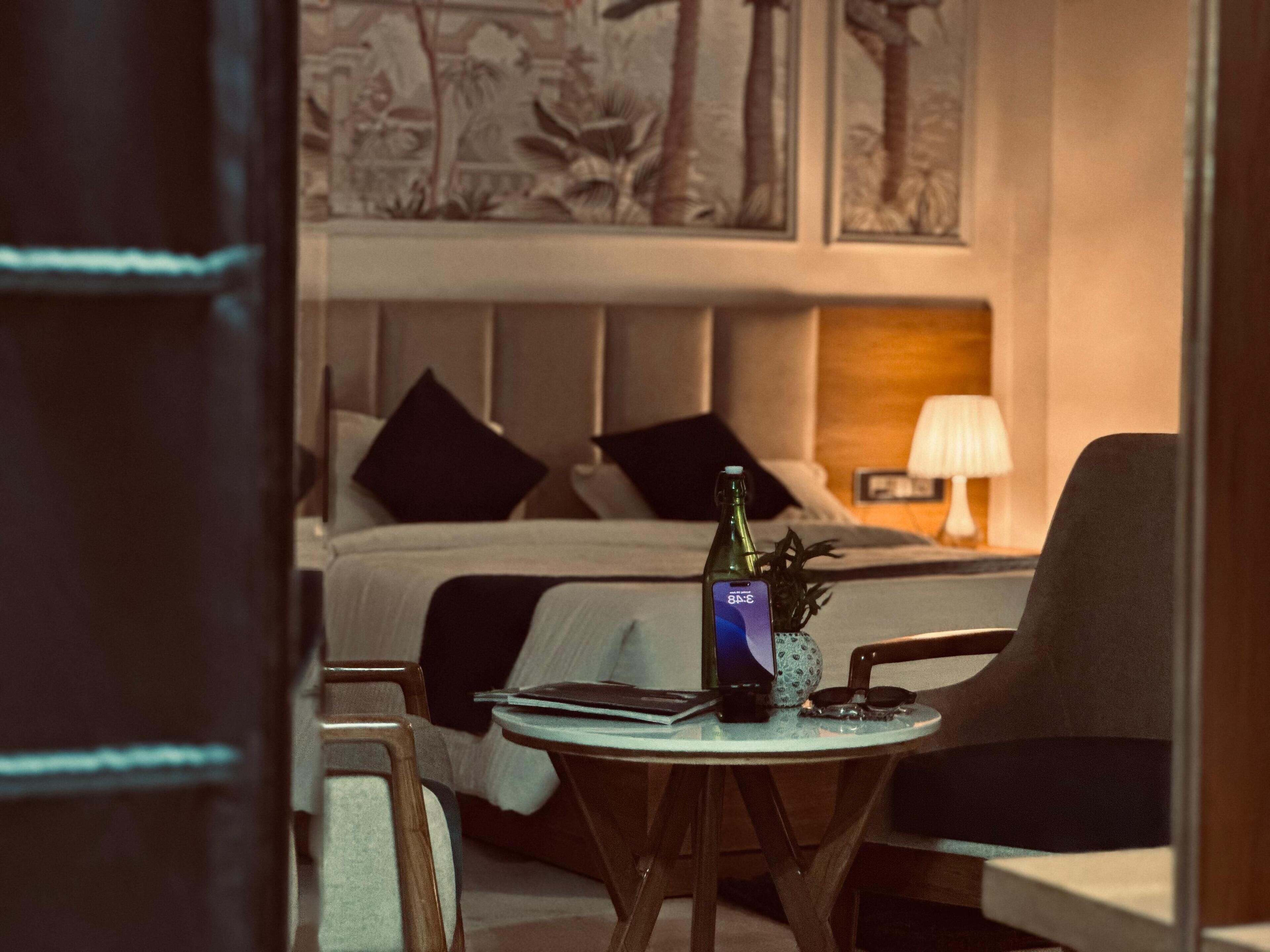 Habitación de hotel con cama, mesita de noche con una botella y vasos, lámpara y una silla.