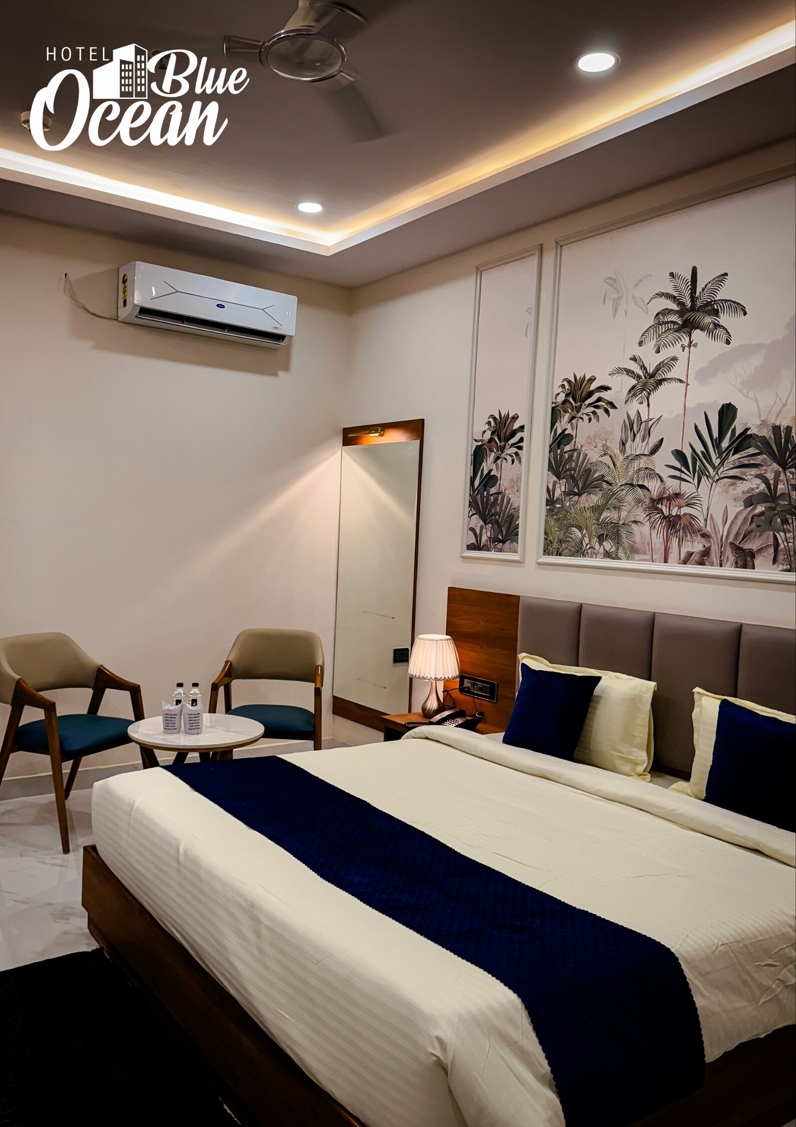 A hotel room with a bed, two chairs, a small table with water bottles, a lamp, and a wall-mounted air conditioner.