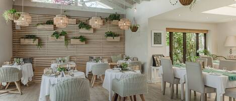 A dining area with multiple tables and wicker chairs, wooden slats on the wall, and hanging planters.