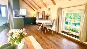 Ein Zimmer mit Holzbalkendecke, einem Esstisch mit Stühlen, einem Fenster mit Vorhängen und einer Küche im Hintergrund.