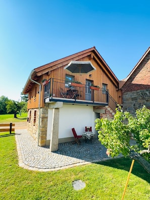 A wooden building with a balcony, a stone wall, and a paved area with a bench.